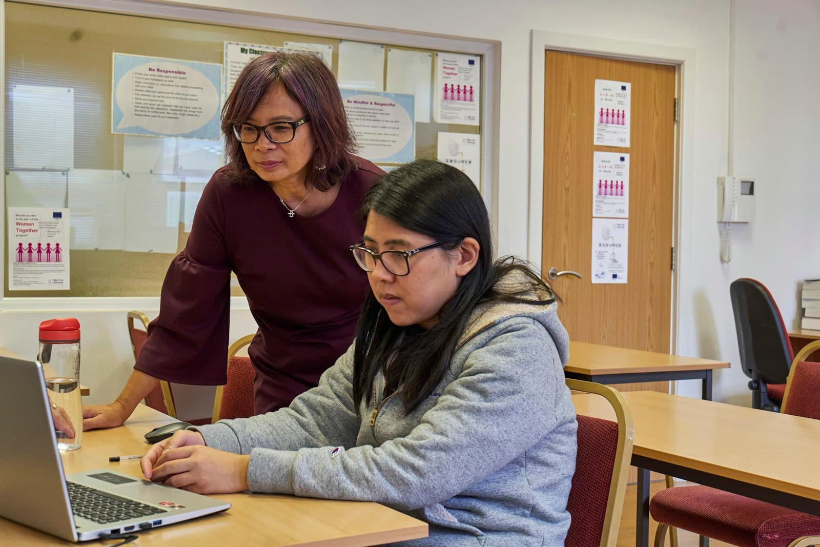 A staff member standing beside a participant, guiding her through a task on a laptop in a community-based workshop setting